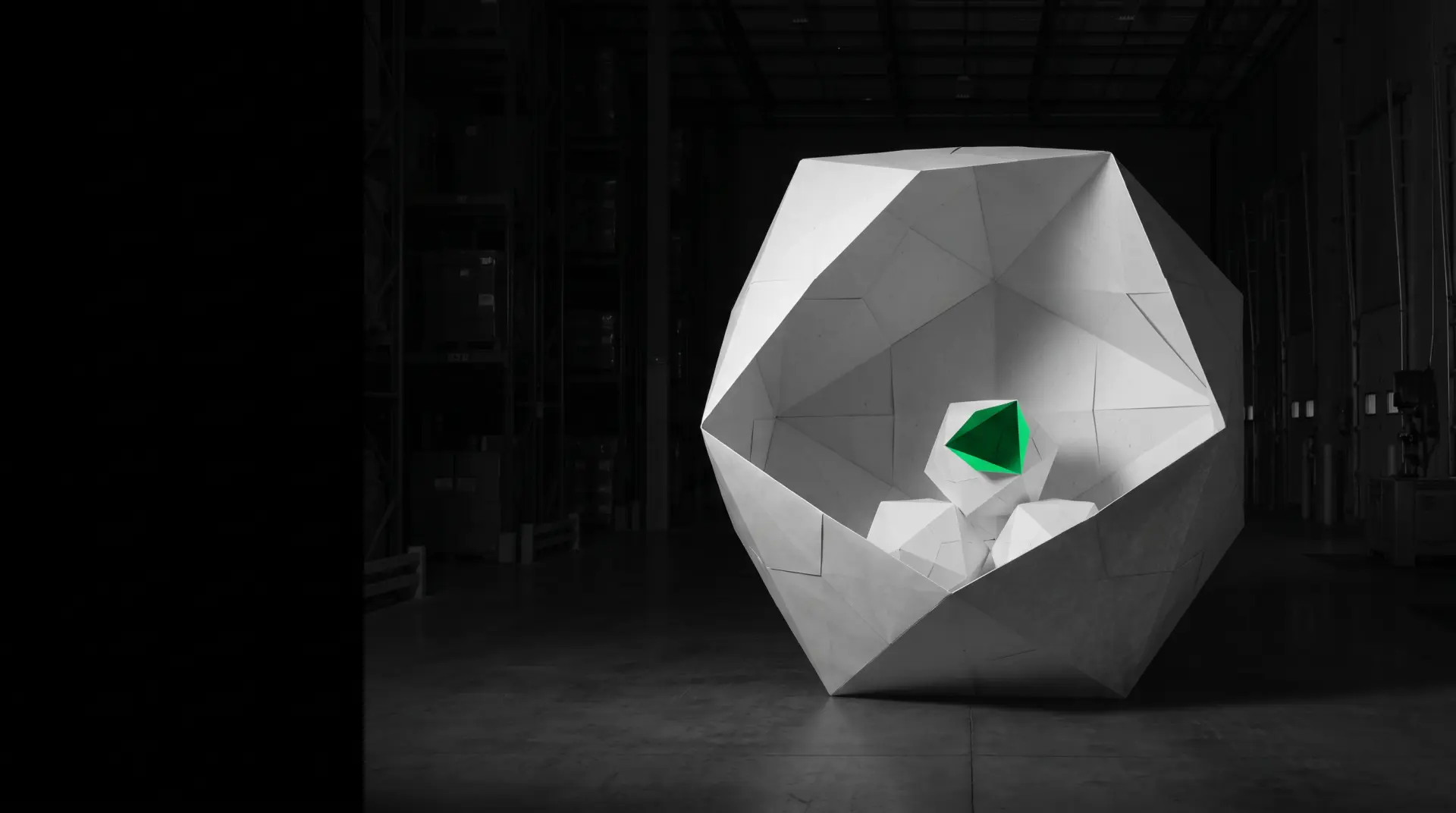 A truck-sized white paper polyhedron opened in section inside a dark warehouse; three small polyhedrons rest inside, only one with a green fold.